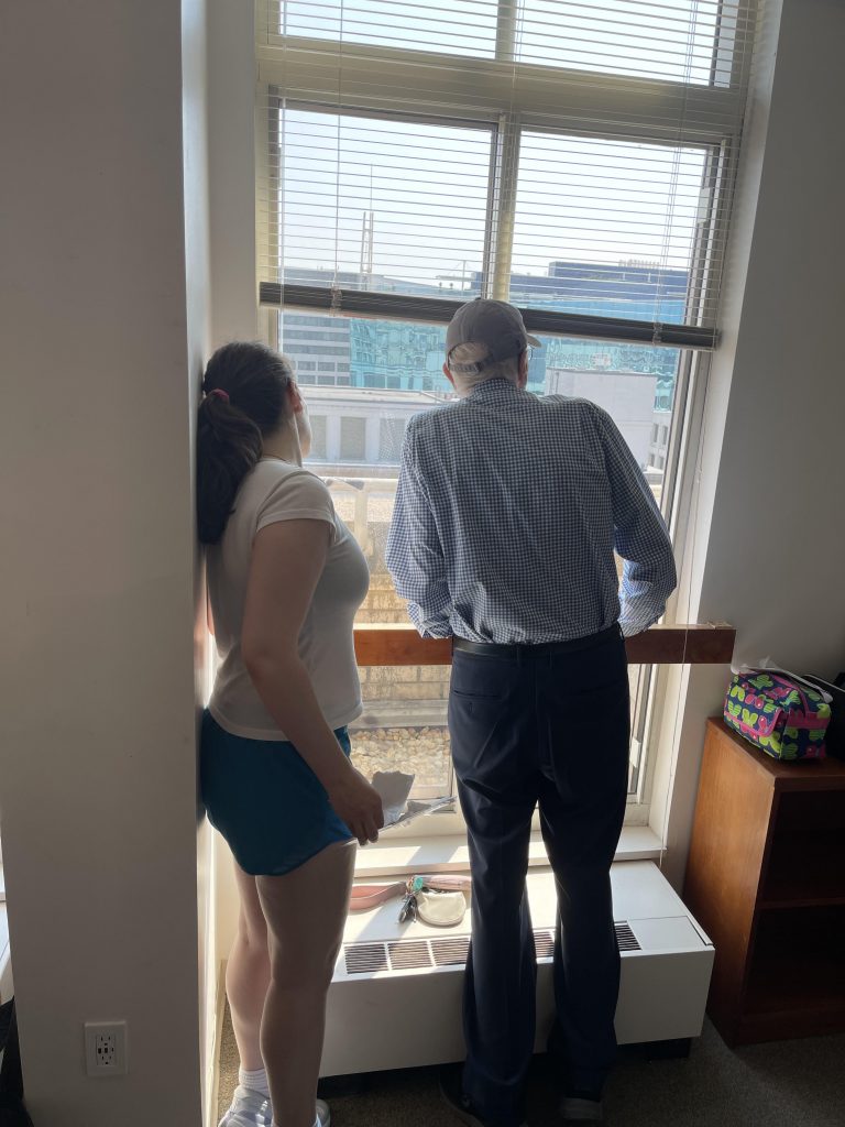 A young woman and her grandfather look out the window of her new Georgetown University dorm room, reflecting an intergenerational moment of transition.